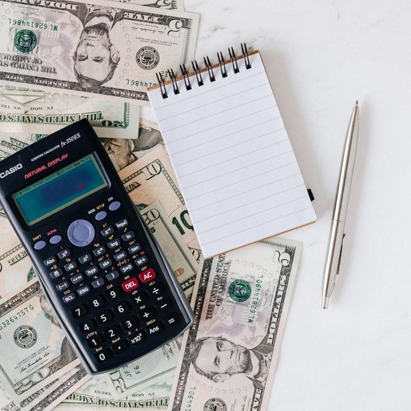 A flat lay image showing US dollar bills scattered on a white surface next to a calculator, a blank spiral notepad, and a silver pen, symbolizing financial planning and investment.