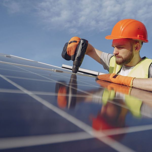 A close-up, upward-angled shot of a solar installer wearing an orange hard hat and safety vest, using a power drill to secure a black solar panel onto a residential roof structure under a blue sky.