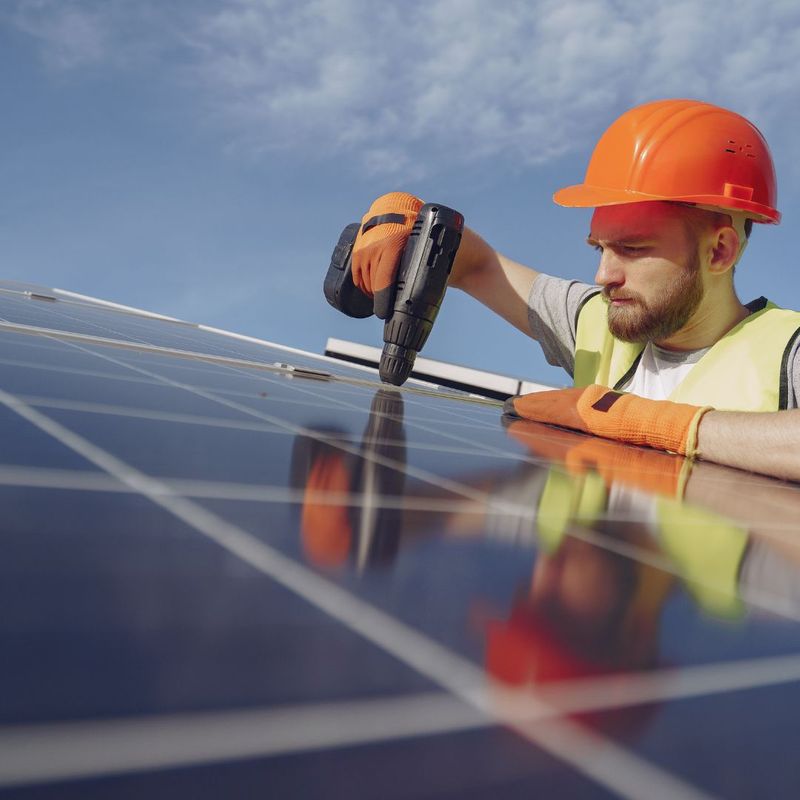 A close-up, upward-angled shot of a solar installer wearing an orange hard hat and safety vest, using a power drill to secure a black solar panel onto a residential roof structure under a blue sky.