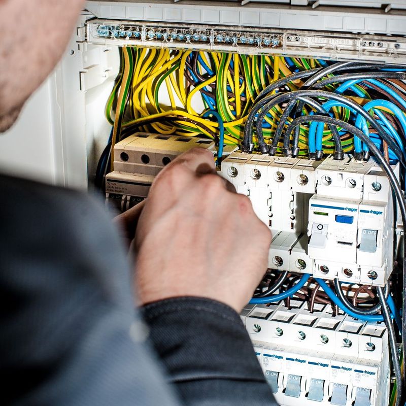 A close-up, over-the-shoulder shot of an electrician's hands expertly working on the colorful wiring inside an open electrical panel.