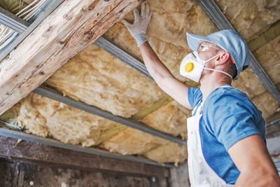 A contractor wearing a respirator mask installs fiberglass insulation in an attic to improve energy efficiency. A contractor wearing a respirator mask installs fiberglass insulation in an attic to improve energy efficiency.