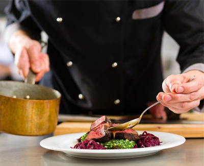 Chef holding a pot, drizzling sauce with a spoon over a meat dish.