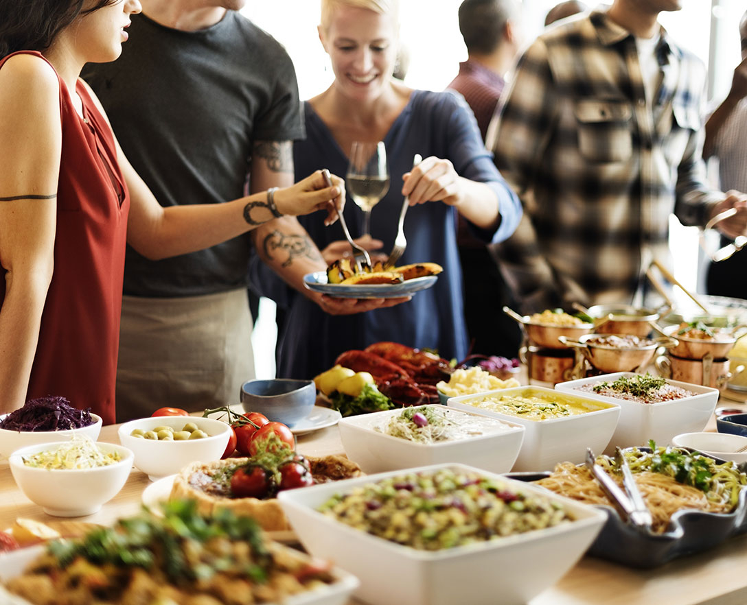 Two women sampling food from a plate being held by a man, standing in line at a large buffet-style food table.