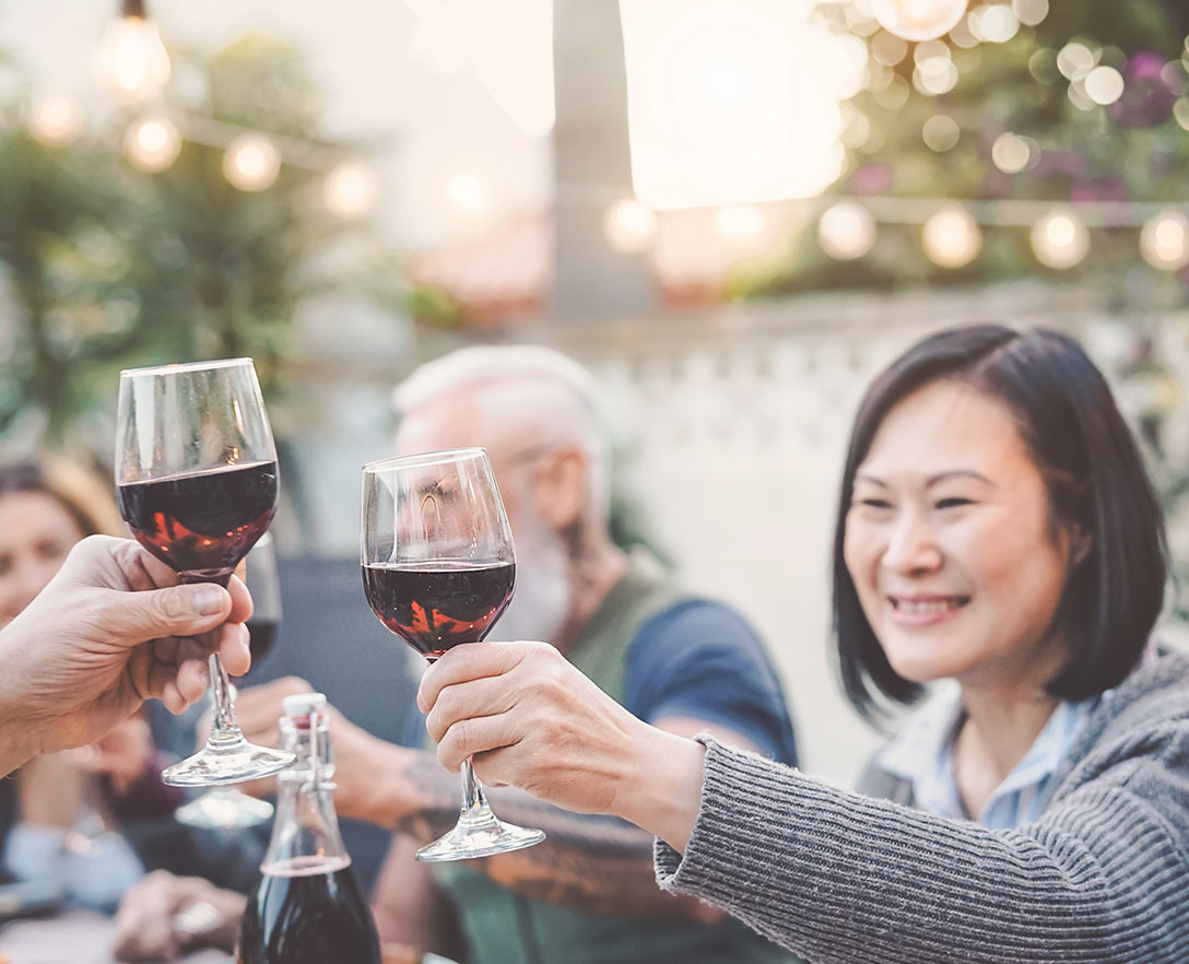 Woman sitting outside with friends holding a glass of red wine clinking, “Cheers” with another glass of red wine.