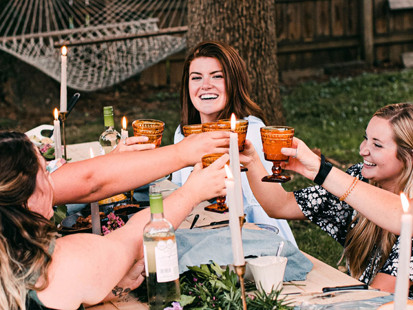 A group of girls sitting around a table, saying cheers with their drinks.