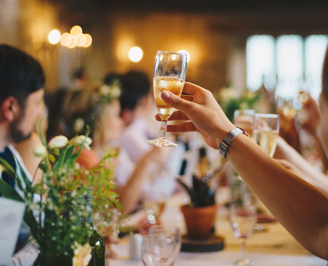 Blurry image of people sitting at a large table, focused on a hand holding up a small champagne flute for a toast.