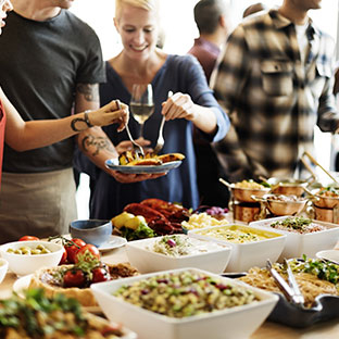 People sampling food from a plate, standing in line at a large buffet-style food table.
