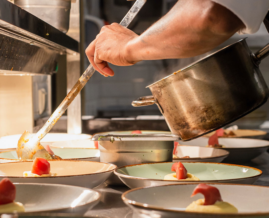  A chef in a kitchen holding a large pot dishing several plates of food with a ladle.