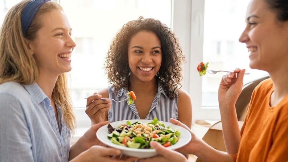 A smiling diverse group of people gathers around a table, lifting a platter of healthy vegetables in a bright, modern setting.