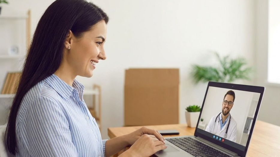 A smiling woman at home connects with a doctor via video conference on her laptop.