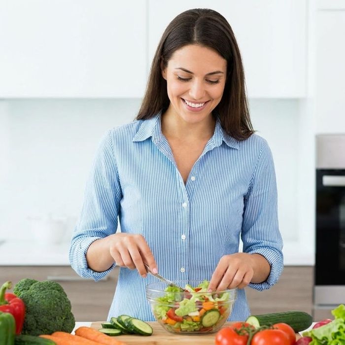 A woman smiles as she prepares a colorful fresh vegetable salad in a bright kitchen.