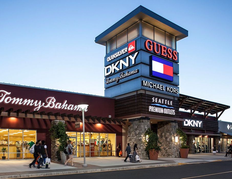 Exterior of a shopping outlet mall with a large sign displaying various store logos, including Quiksilver, Guess, DKNY, Tommy Bahama, Michael Kors, Seattle Premium Outlets, and more. People are walking near the store entrance at dusk.
