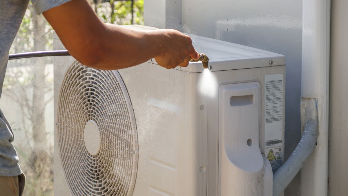 Rinsing the outdoor condenser unit of a residential air conditioner with a water hose during routine AC maintenance in Greenacres, FL