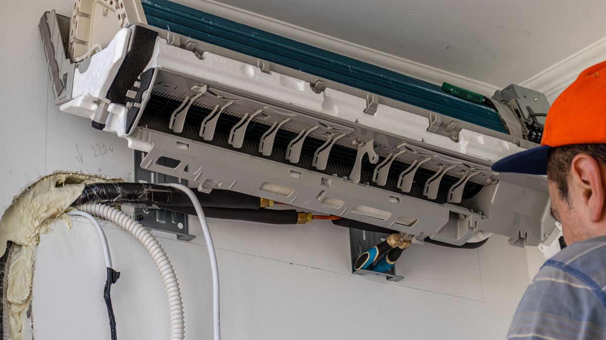 Technician servicing a split AC indoor unit, with refrigerant lines and drain hose visible