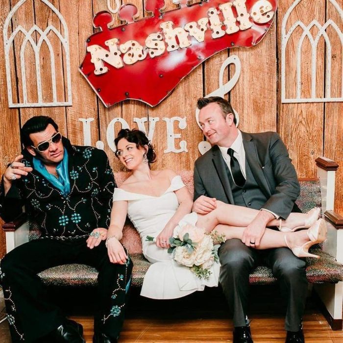A newlywed couple poses with an Elvis impersonator on a bench in front of a rustic Nashville backdrop.