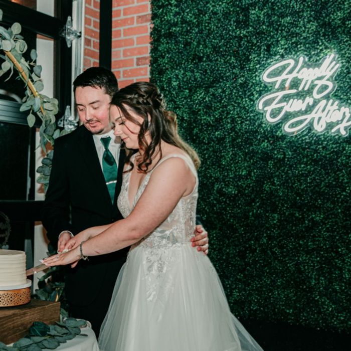 A bride and groom smile while cutting their wedding cake together in front of a greenery wall with a neon sign.