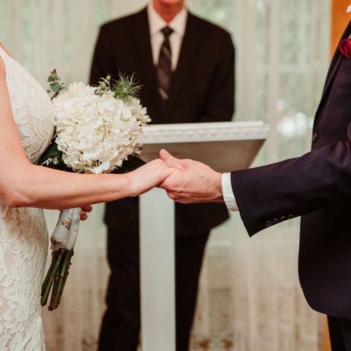 A bride and groom hold hands during their intimate wedding ceremony at a podium with a white floral bouquet.