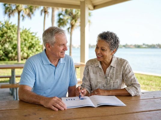 A smiling senior man and woman review charts in a document at a picnic table, with a waterfront and palm trees in the background. Senior Couple Discussing Documents Outdoors