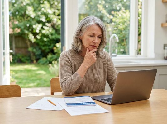A thoughtful senior woman reviews information on her laptop. Documents, including 'Medicare and You,' are on the table in a sunny room. Senior woman researching health information on laptop