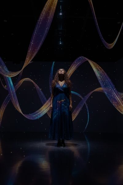 A woman looks up, captivated by photorealistic, shimmering ribbons of light that dance and intertwine, visually interpreting the melody of a cello in a synesthesia room.