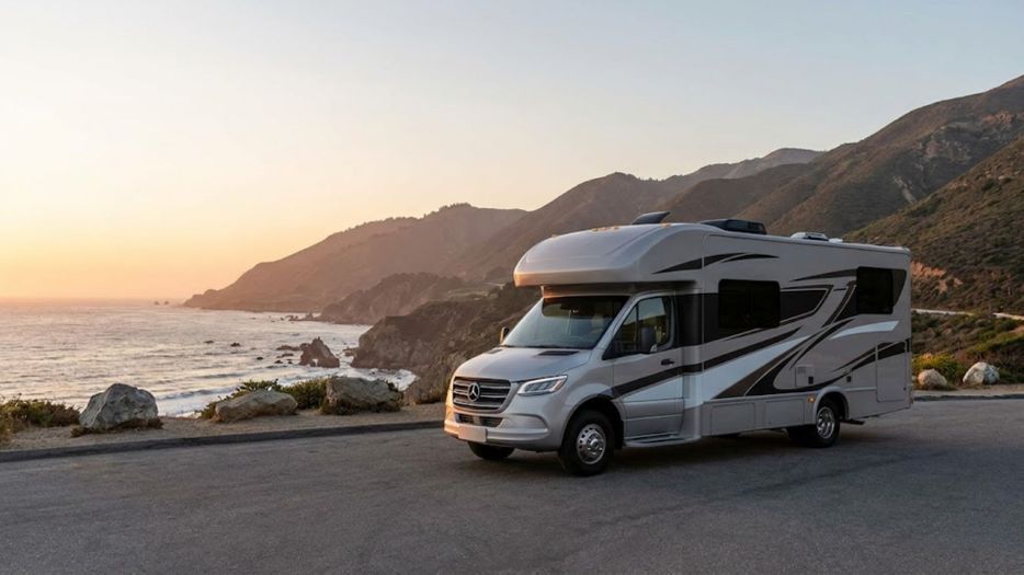 A large, modern recreational vehicle is parked near a California coastline with mountains during a serene sunset.