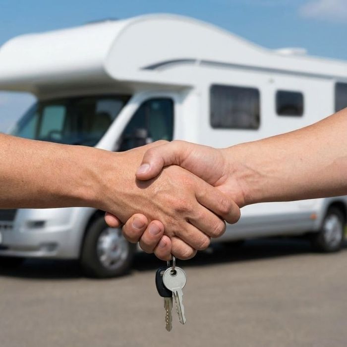 Two hands shake over a set of RV keys in the foreground, with a white motorhome blurred in the background, representing a successful sale.