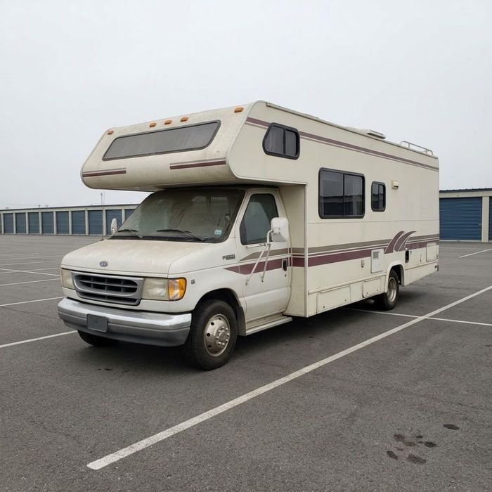 A dusty, older recreational vehicle sits unused in a large, fenced storage lot.