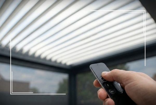 Hand holding a remote control to operate a louvered patio roof, which is opening in the background.