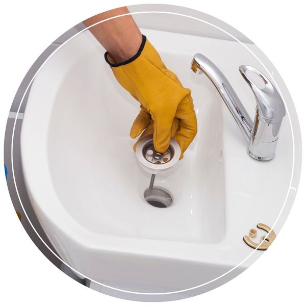 A hand wearing a yellow leather work glove holding a metal sink strainer over the drain opening of a white porcelain bathroom sink.