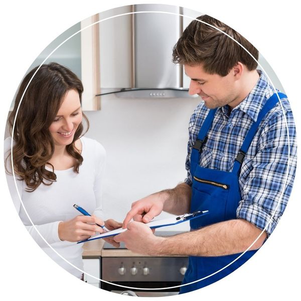 A smiling woman in a kitchen signing a clipboard held by a male plumber wearing blue coveralls and a plaid shirt.