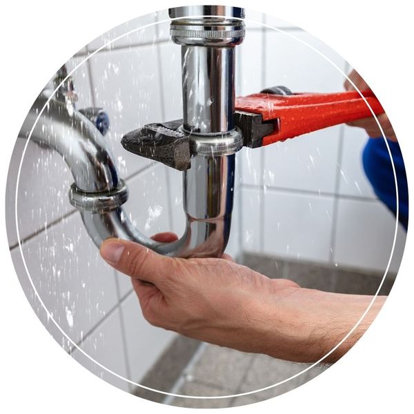 A plumber using a large red pipe wrench to tighten a leaking, water-spraying connection on a chrome P-trap pipe.