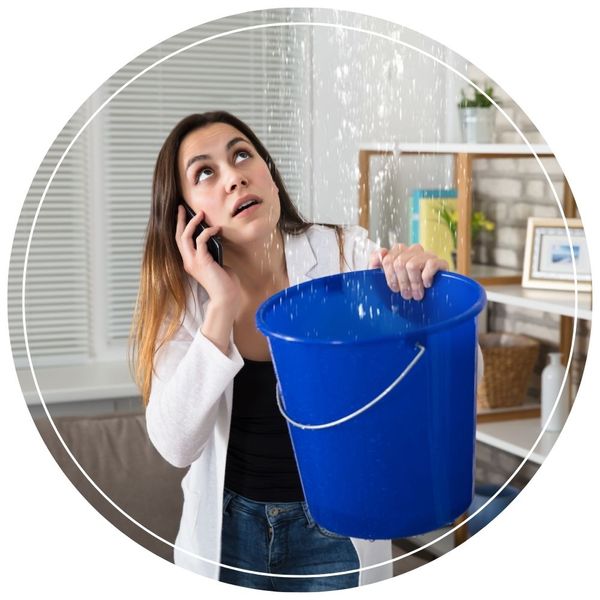 A worried woman holding a blue bucket while talking on the phone, catching a severe water leak dripping from the ceiling.