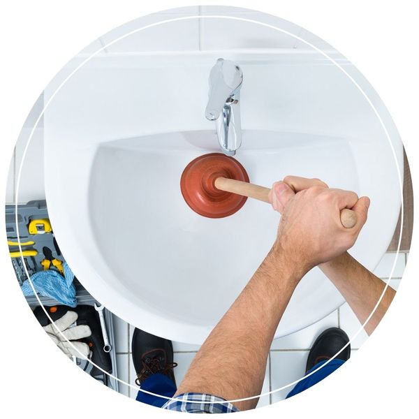 Top-down view of a person using a wooden-handled plunger to clear a blockage in a white bathroom sink, with a gray toolbox on the floor nearby.