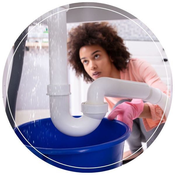 Close-up of a leaking white plastic P-trap assembly dripping water into a blue bucket beneath a sink, with a concerned woman looking on in the background.