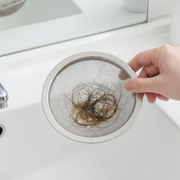 A hand holds a metal sink strainer filled with collected hair and debris over a clean white bathroom sink.