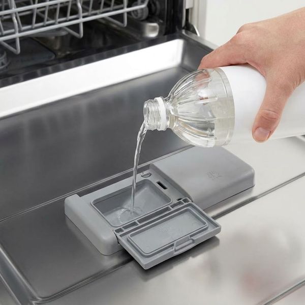 A hand holds a plain bottle and pours clear white vinegar into the open detergent compartment of a dishwasher. A hand holds a plain bottle and pours clear white vinegar into the open detergent compartment of a dishwasher.