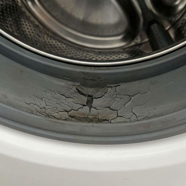 A detailed macro photograph shows a severely cracked and degraded rubber door seal on a front-loading washing machine. A detailed macro photograph shows a severely cracked and degraded rubber door seal on a front-loading washing machine.