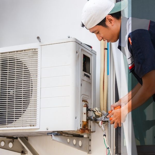 HVAC specialist in a white cap performing maintenance on an outdoor mini-split condenser unit, checking refrigerant lines for peak winter performance.