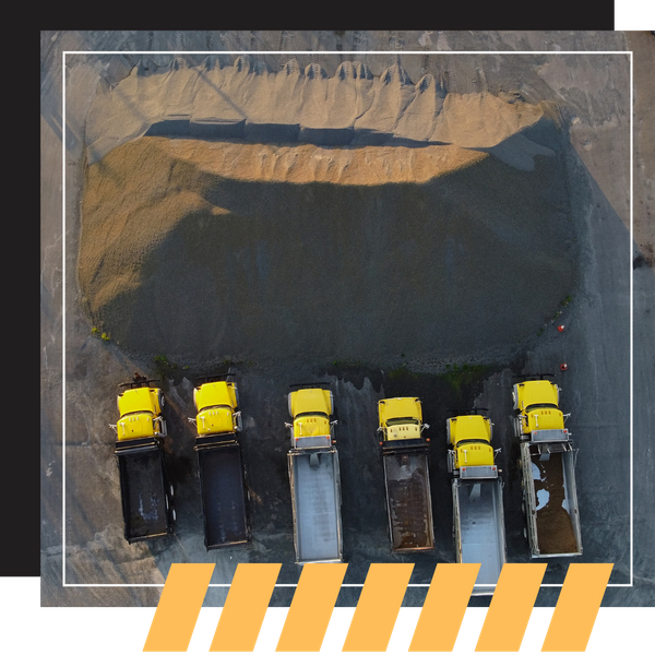 An aerial view of six yellow dump trucks lined up at the base of a massive gravel stockpile.