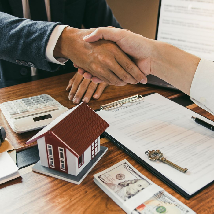 Shaking hands over a home contractor, with a key, model home, and stack of cash on desk