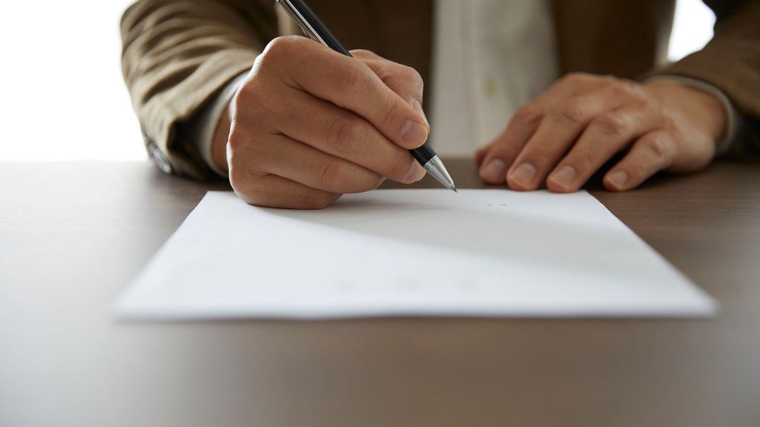 A close-up of a person's hands in a professional suit signing a legal document with a pen on a wooden desk. A close-up of a person's hands in a professional suit signing a legal document with a pen on a wooden desk.