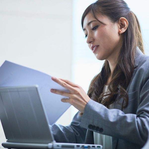 A professional woman in a grey suit reviews paperwork while working on a laptop in a bright office setting.