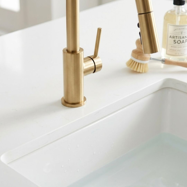a modern brushed gold kitchen faucet with a pull-down sprayer and a white farmhouse sink on a quartz island
