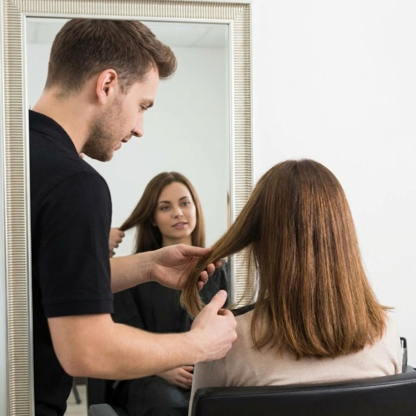 A male hairstylist and a female client sitting in front of a mirror, consulting about her hair's suitability for a perm