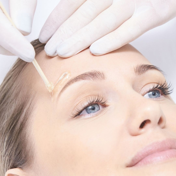 Gloved hand applies wax above woman's eyebrow with a small stick.