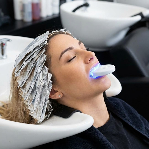 A woman is relaxing in a salon shampoo bowl with hair color processing foils and a blue light teeth whitening tray in her mouth
