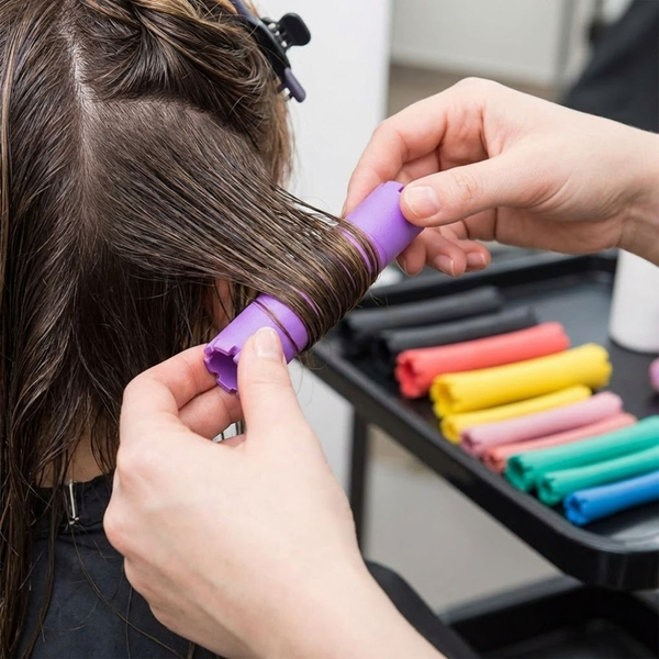 a hairstylist's hands wrapping hair around a large, purple perm rod, with other colorful rods visible, demonstrating the modern approach to perming