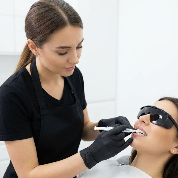 A salon professional in a black apron is carefully applying a professional teeth whitening gel to a client's teeth