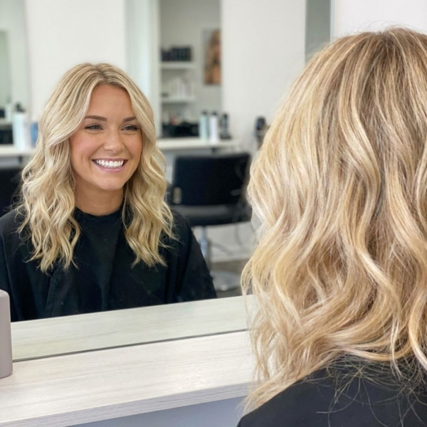 A woman with freshly styled blonde hair is smiling confidently at her reflection in a salon mirror, showing off her bright white teeth