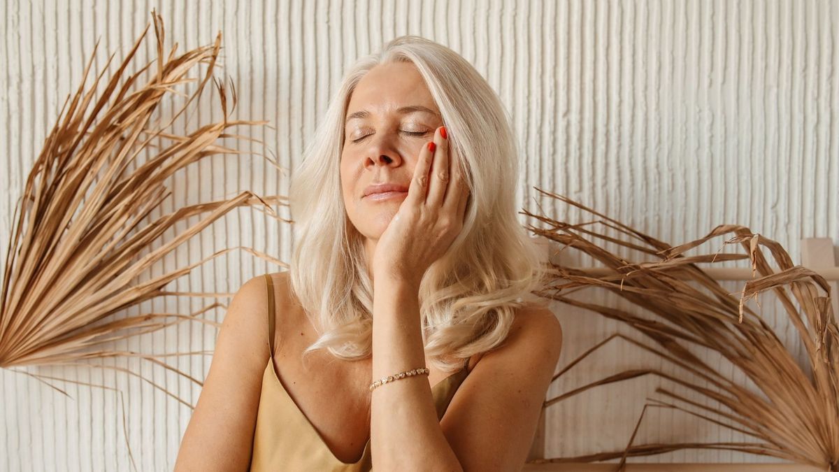 A serene woman with blonde hair and eyes closed, gently touching her smooth cheek.